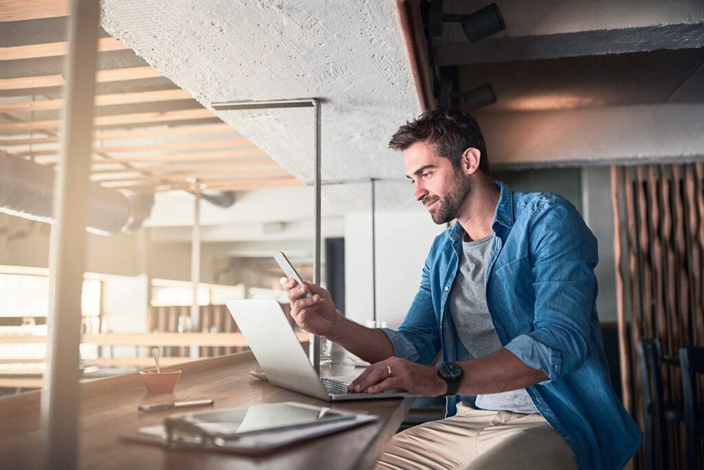 Homem visualizando um celular em uma mesa de escritório junto a um notebook